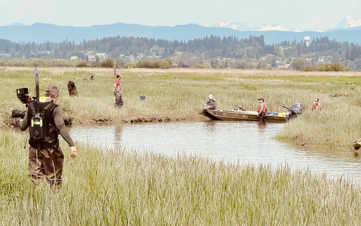 A camera operator and film crew working beside a small boat in a tidal marsh, with wooded hills and snowy peaks in the distance